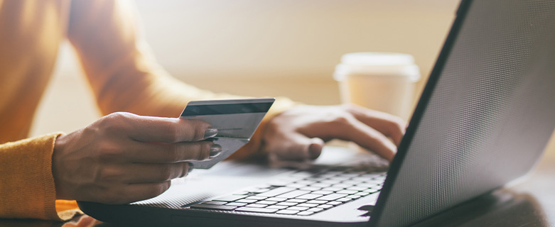 a person holds a credit card with one hand and types on a laptop with the other, a cup of coffee is in the background